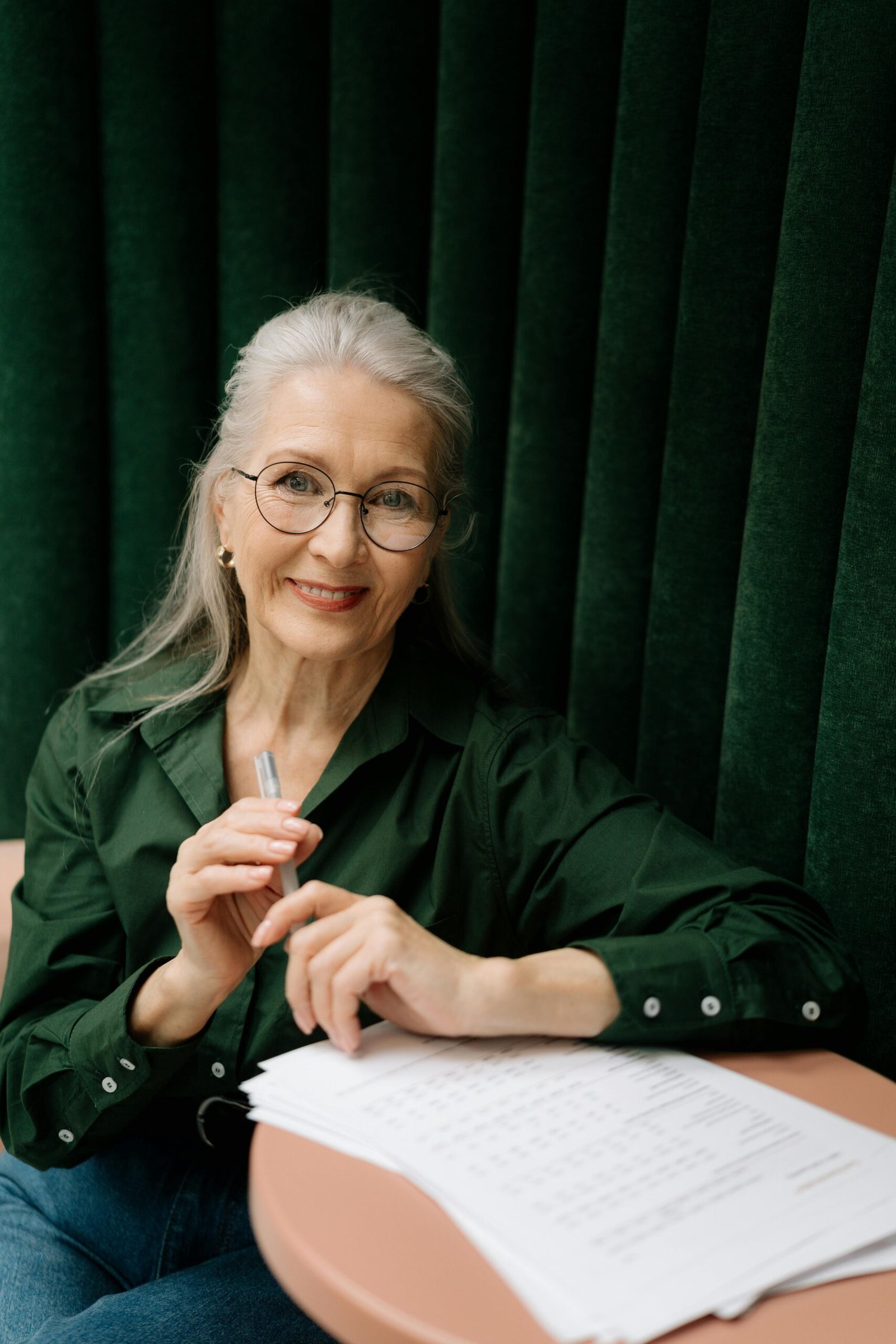 Elegant senior woman with eyeglasses smiling while holding a pen and reviewing documents indoors.