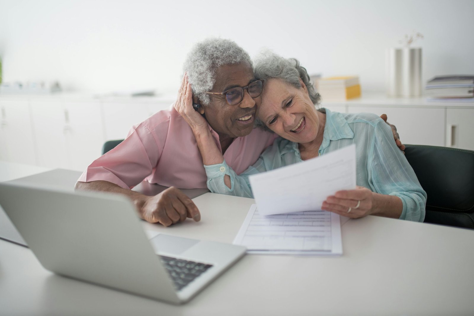 A joyful senior couple smiling while reviewing documents together at a desk with a laptop.