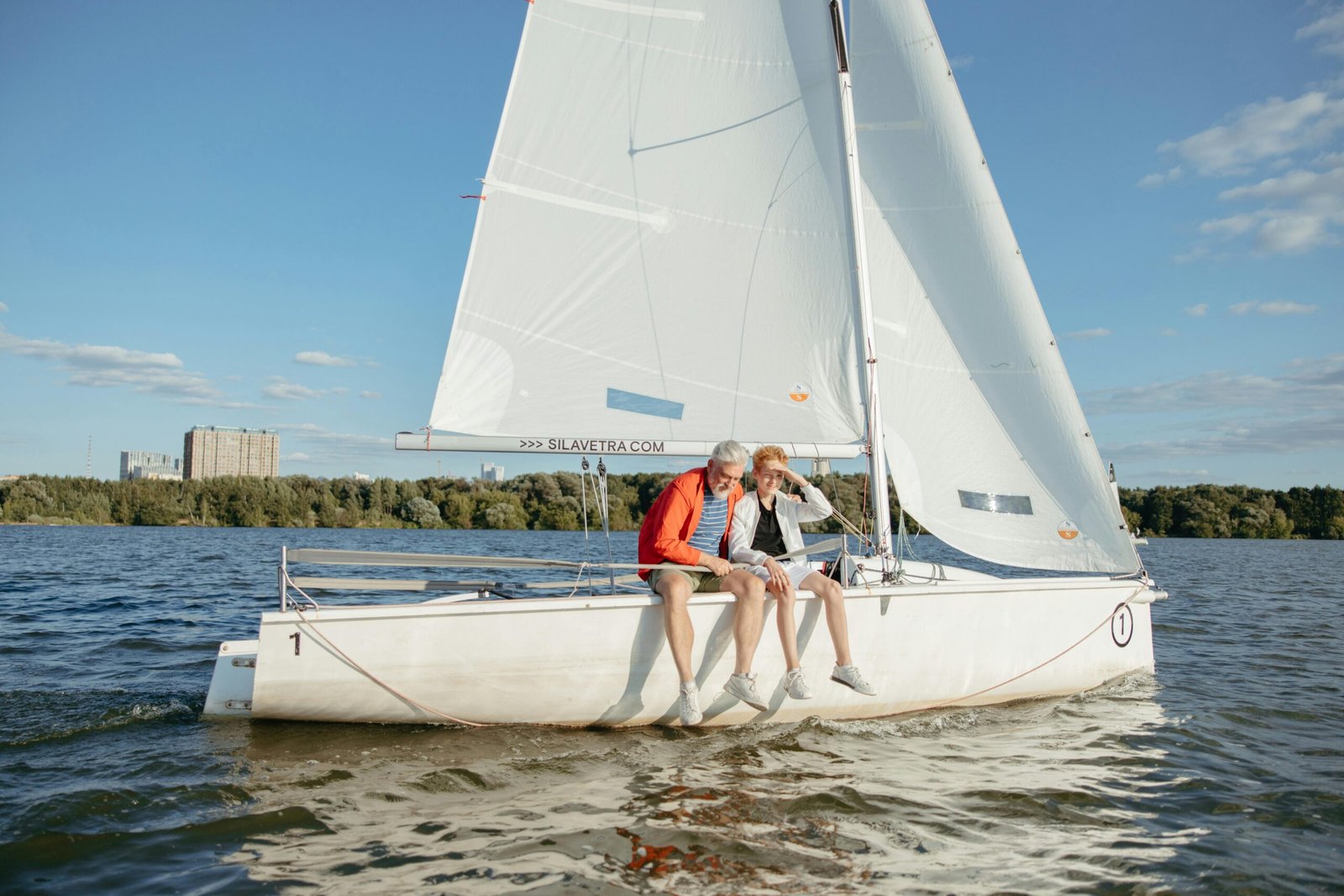 Grandfather bonds with grandson while sailing a white boat on a sunny lake day.